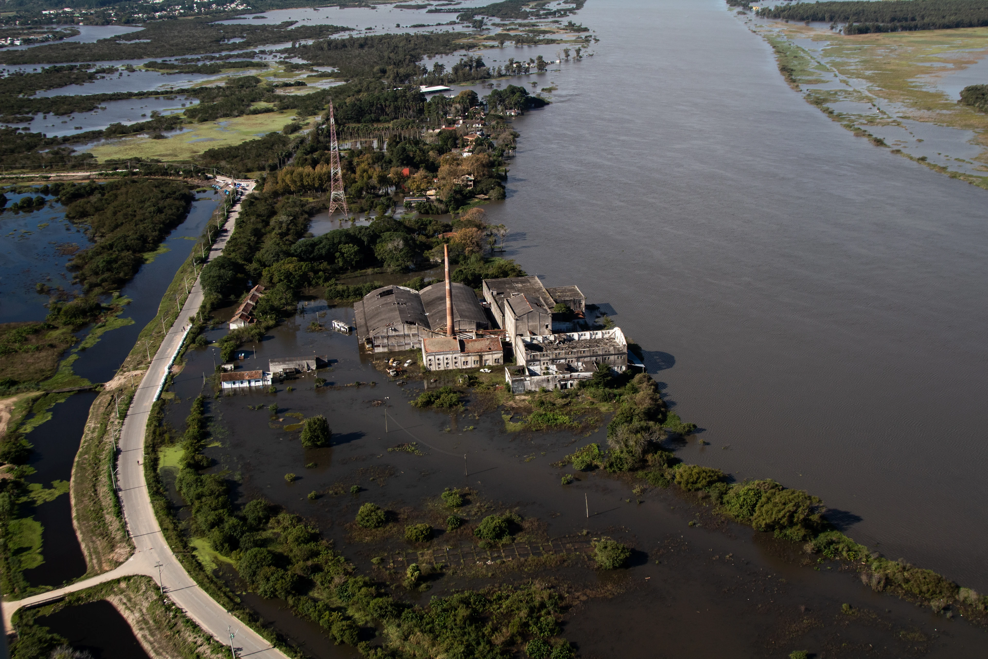 Old industrial buildings and a tall chimney surrounded by floodwater beside a wide river