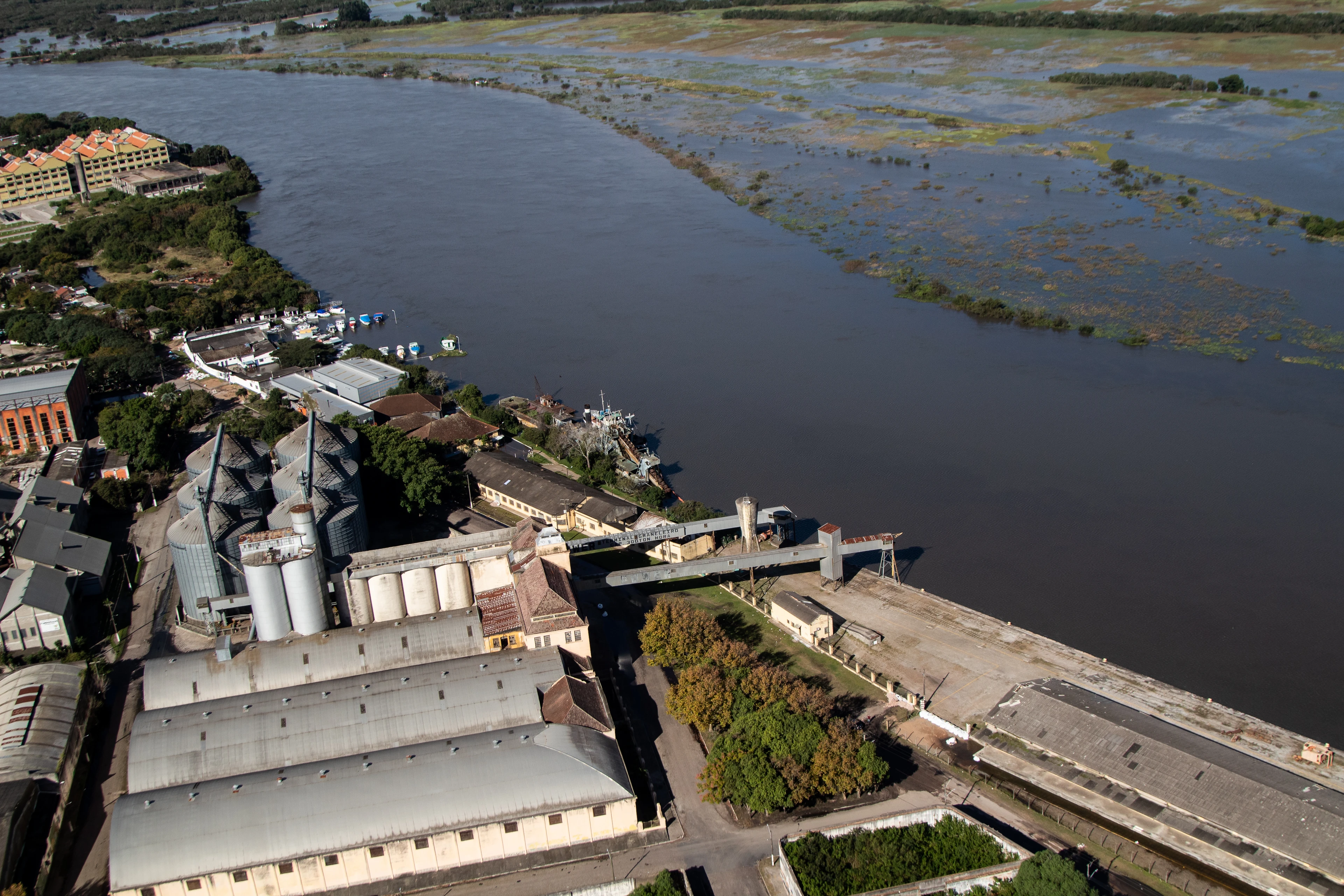 Grain silos, warehouses and a quay beside a swollen river, with flooded wetland on the far bank